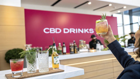 a woman holding a cocktail glass with an orange liquid at a bar and a pink wall reading 'CBD Drinks' in the background