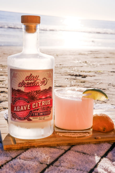 Bottle of Day Dream Agave Citrus THC mixer with a cocktail on a wooden coaster by the beach.